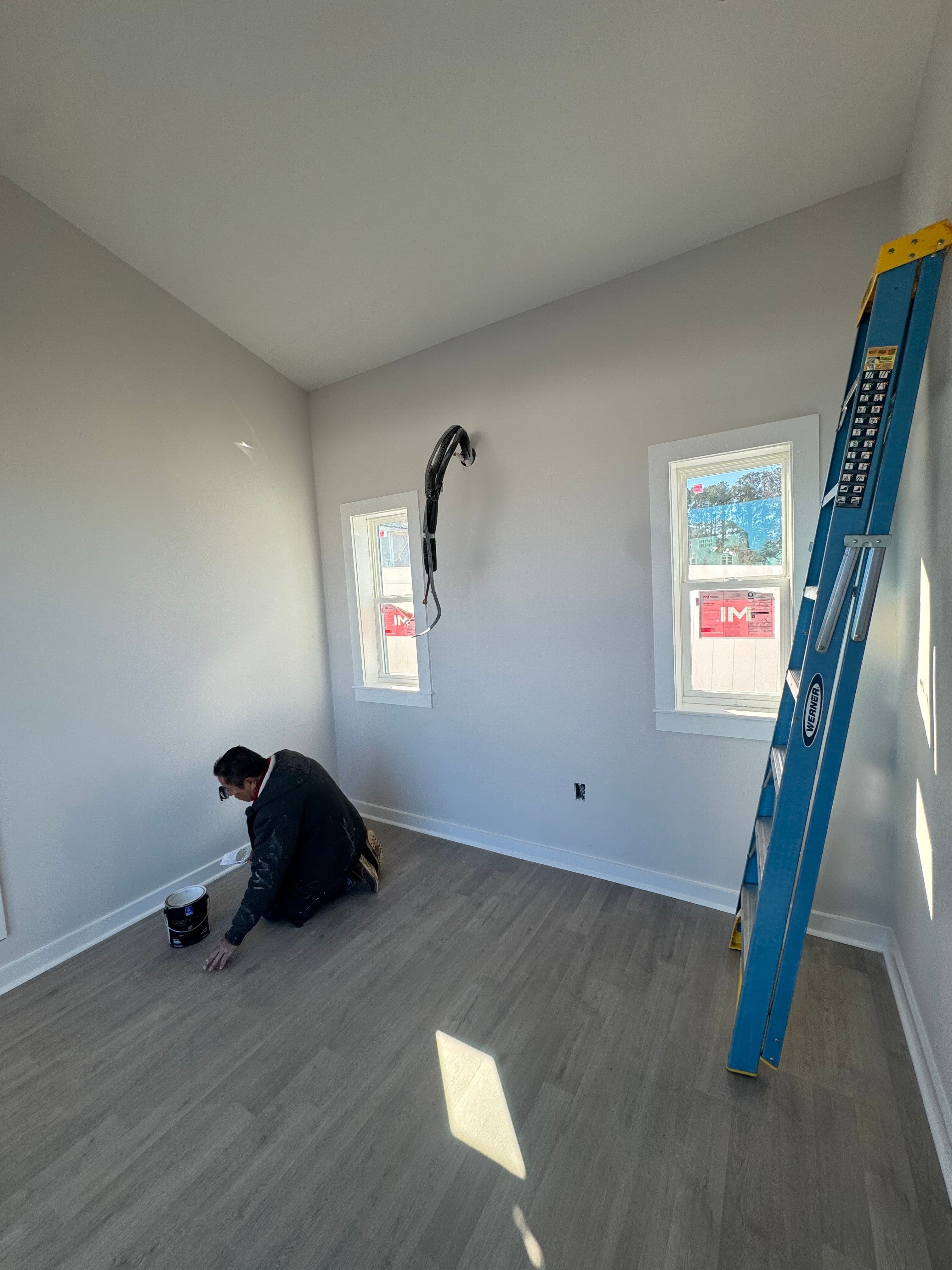 Man painting a wall in a bright room with windows and a ladder.